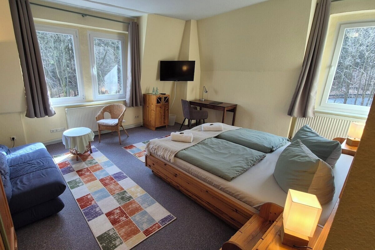 A bedroom with a bed, a chair, a desk, a television, and large windows.