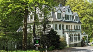 A large, white building with multiple windows and a balcony, surrounded by lush greenery. - Naturhotel Forsthaus (Annaberg-Buchholz)