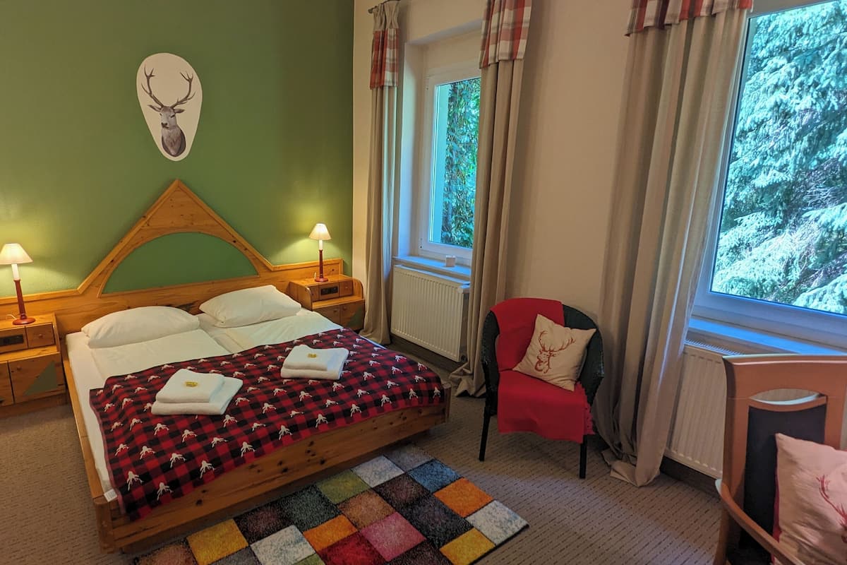A cozy bedroom with a wooden bed, a red and black checkered blanket, a deer head wall decoration, and a view of trees through the window.