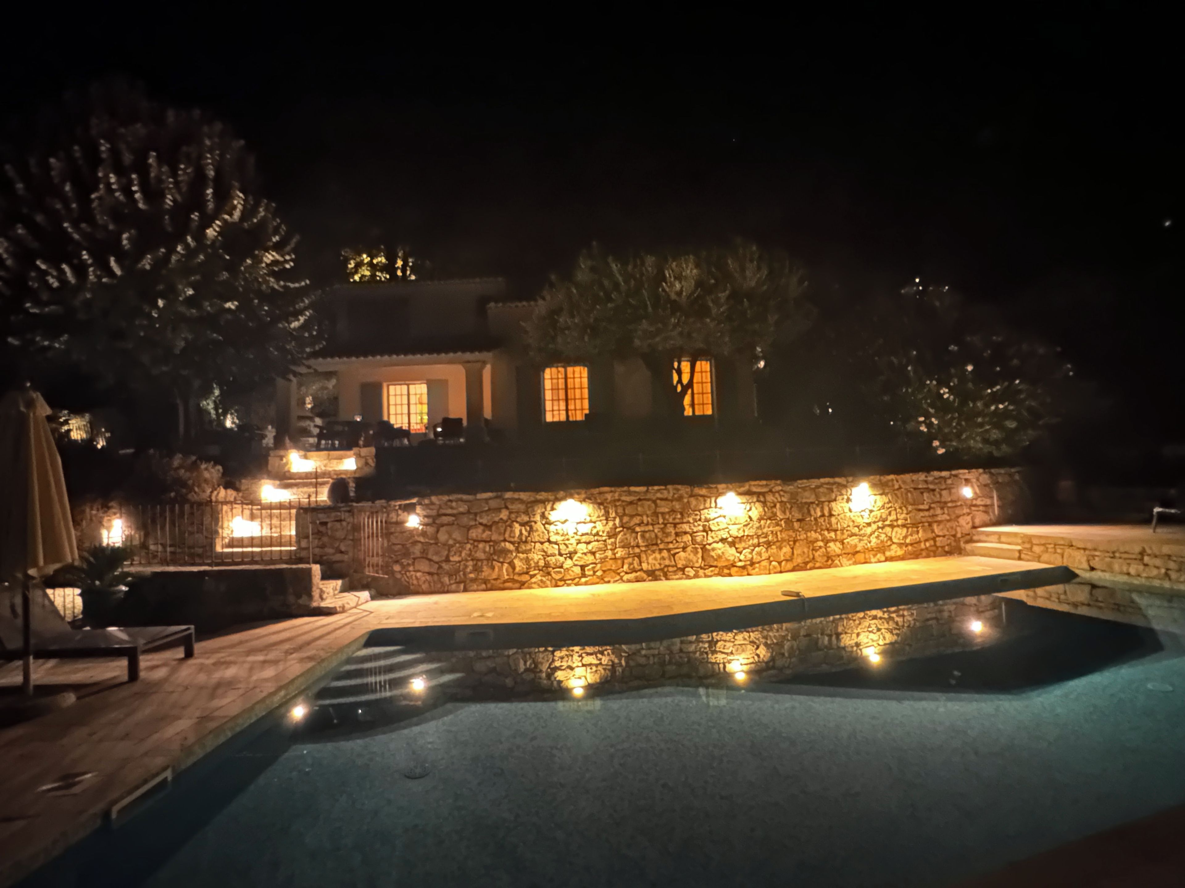 Vista nocturna de un área de piscina con muros de piedra y una terraza.
