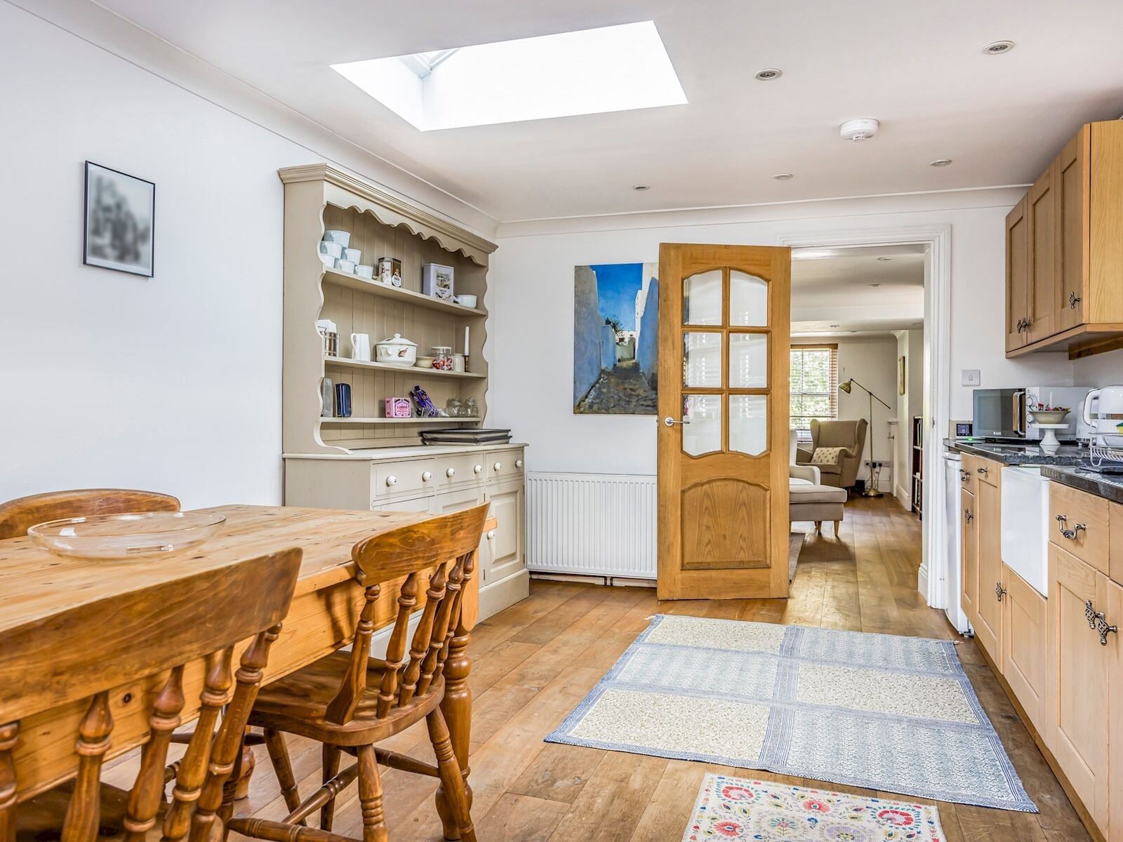 A kitchen with wooden cabinets, a dining table, and chairs. There is a skylight above the kitchen, and a painting on the wall.
