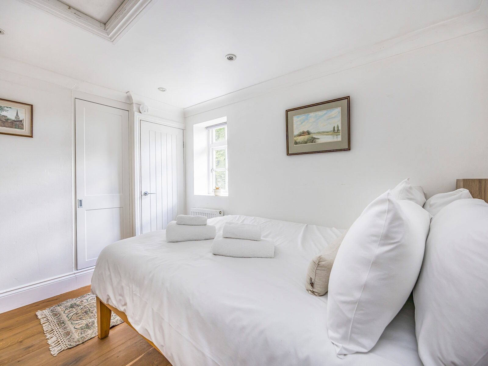 A bedroom with a large bed, white bedding, a wooden floor, a radiator, and a window with a view of greenery.