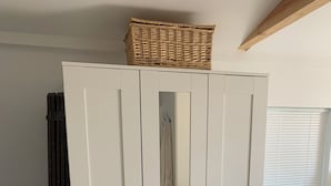 En vit garderob med en flätad korg ovanpå, en svart stol och ett fönster med persienner.