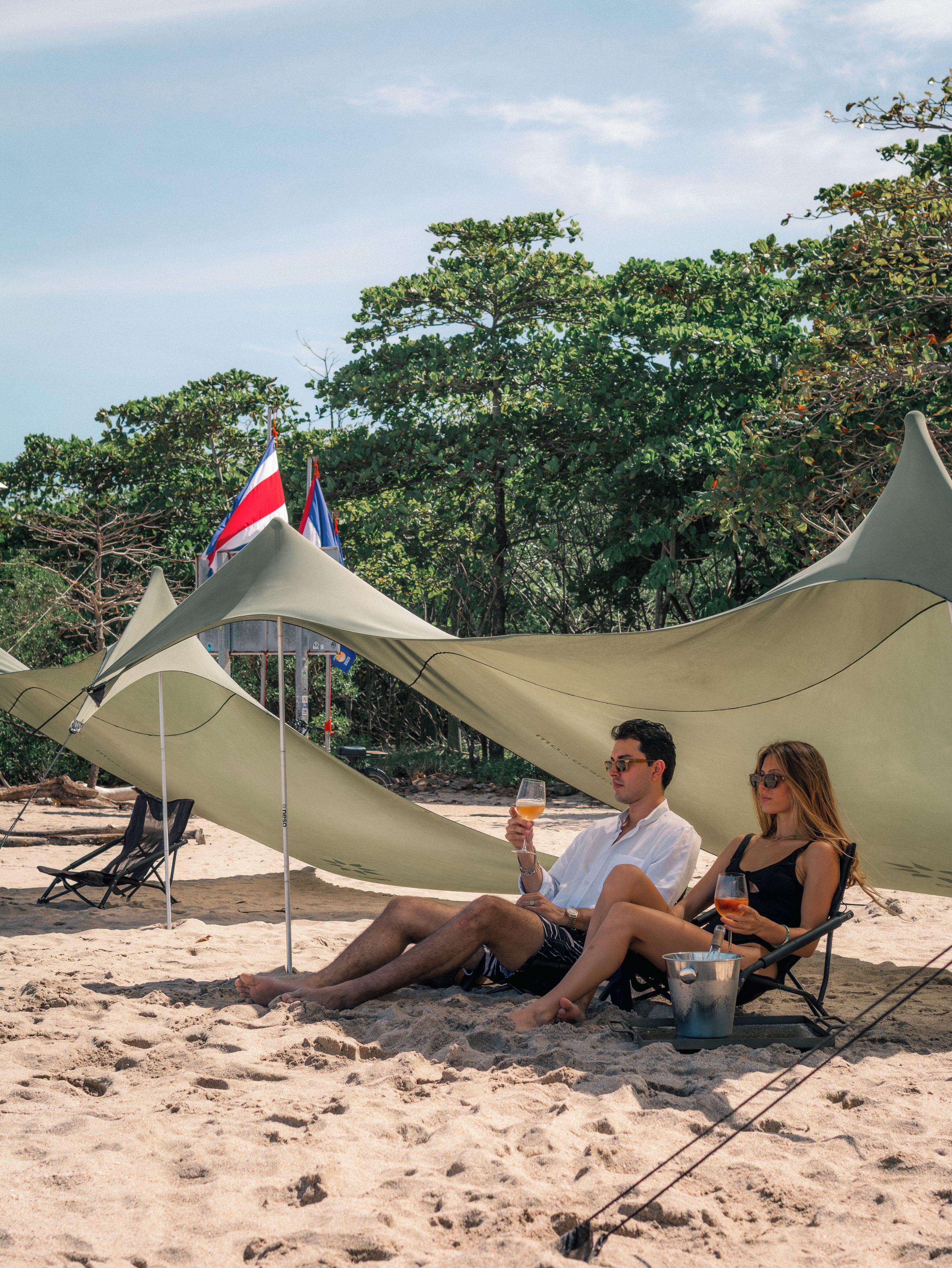 Une scène de plage où un couple est assis sous une tente, avec un drapeau et un ciel dégagé.