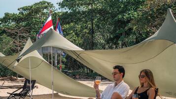 Une scène de plage où un couple est assis sous une tente, avec un drapeau et un ciel dégagé.