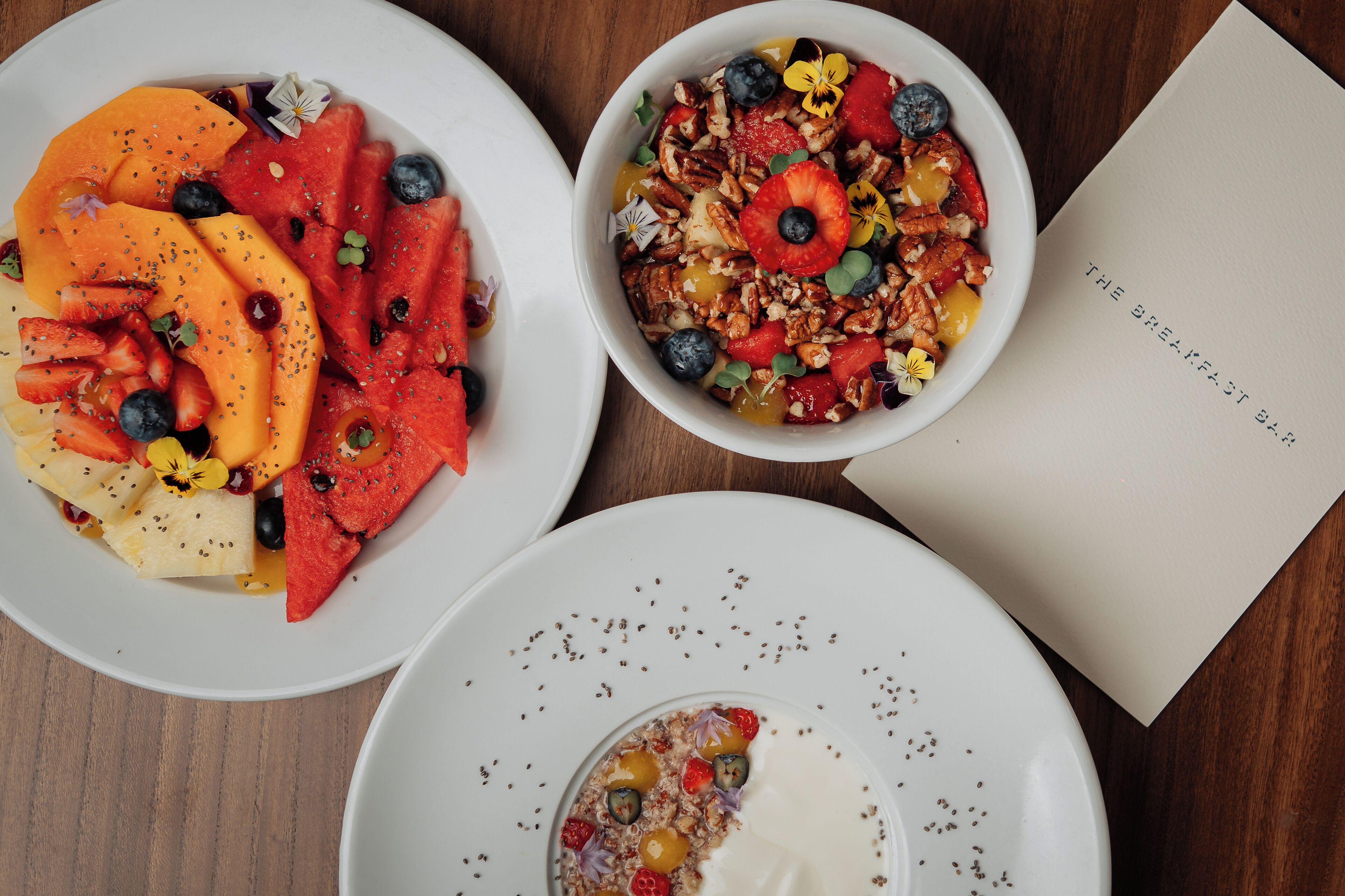 Three bowls of fruit and granola on a wooden table with a menu.