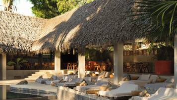 A poolside lounge area with thatched roofs, sun loungers, and a dining area.