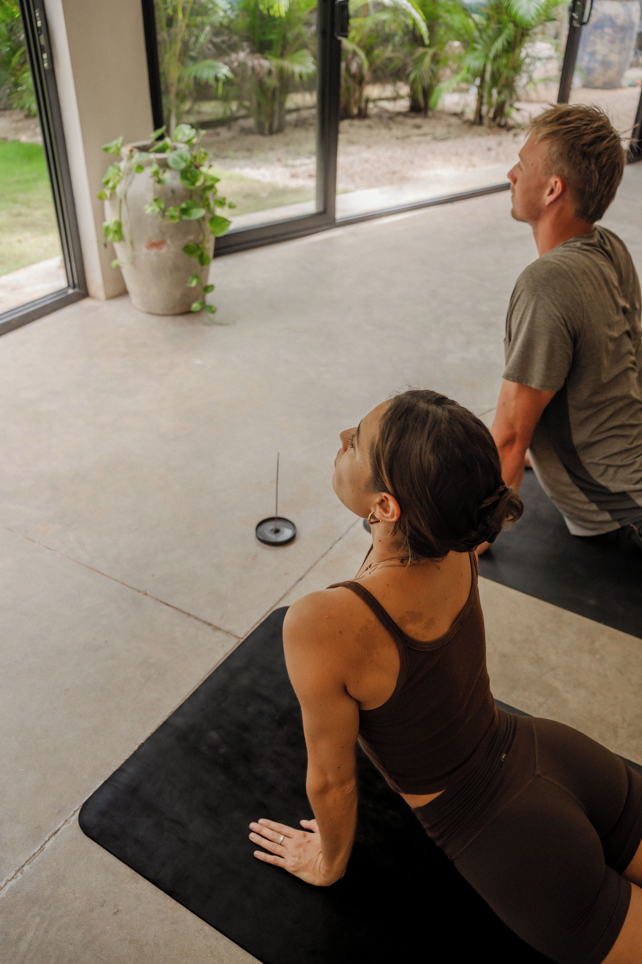A person performing a yoga pose on a black mat in a room with a large window, a potted plant, and a concrete floor.