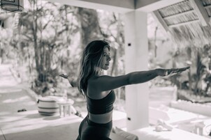 A person in a sports bra and leggings performing a yoga pose outdoors, with a thatched roof structure and a pool in the background.