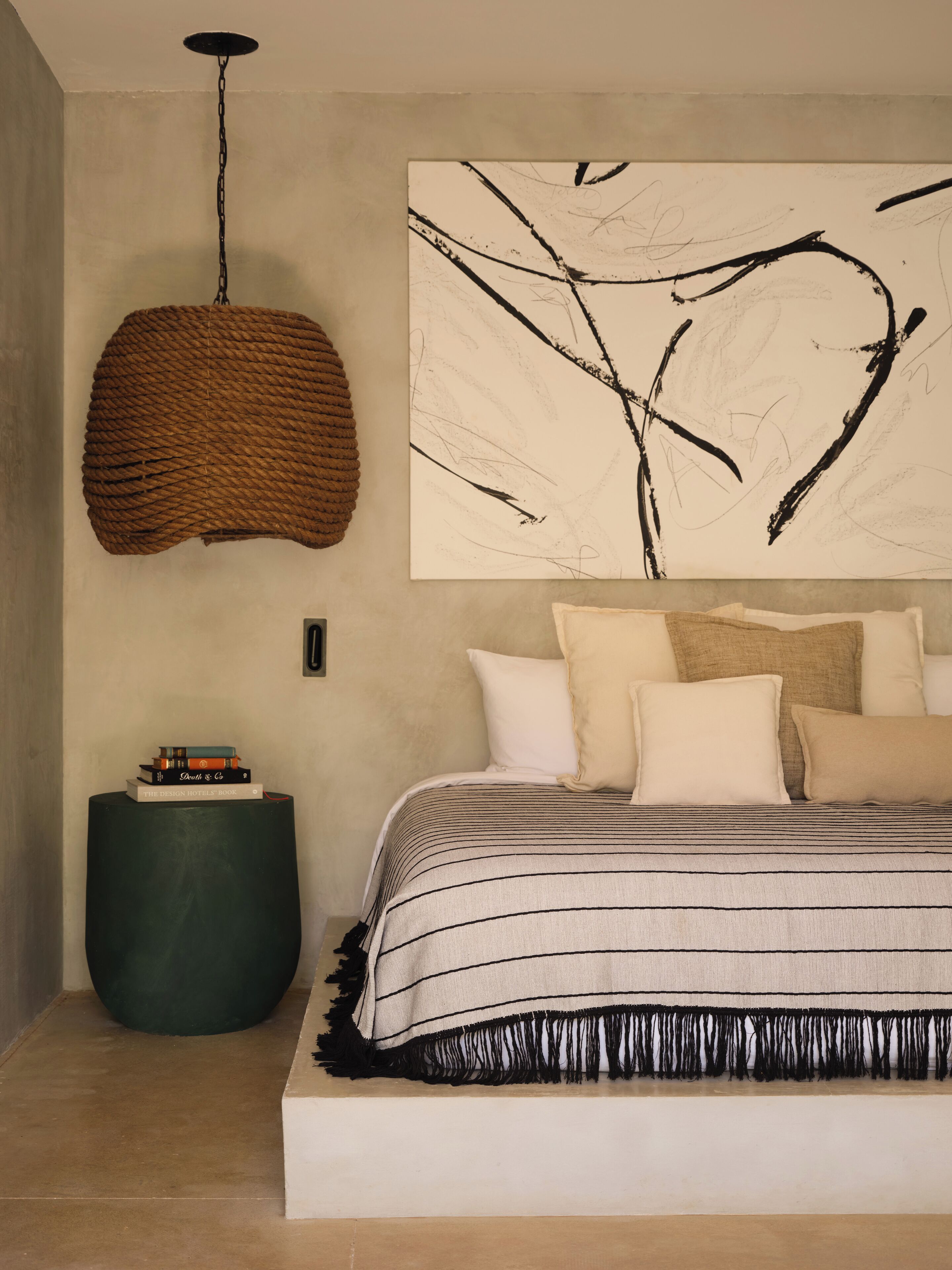 A bedroom with a large bed, a hanging light fixture, a wall art piece, and a small side table with books.