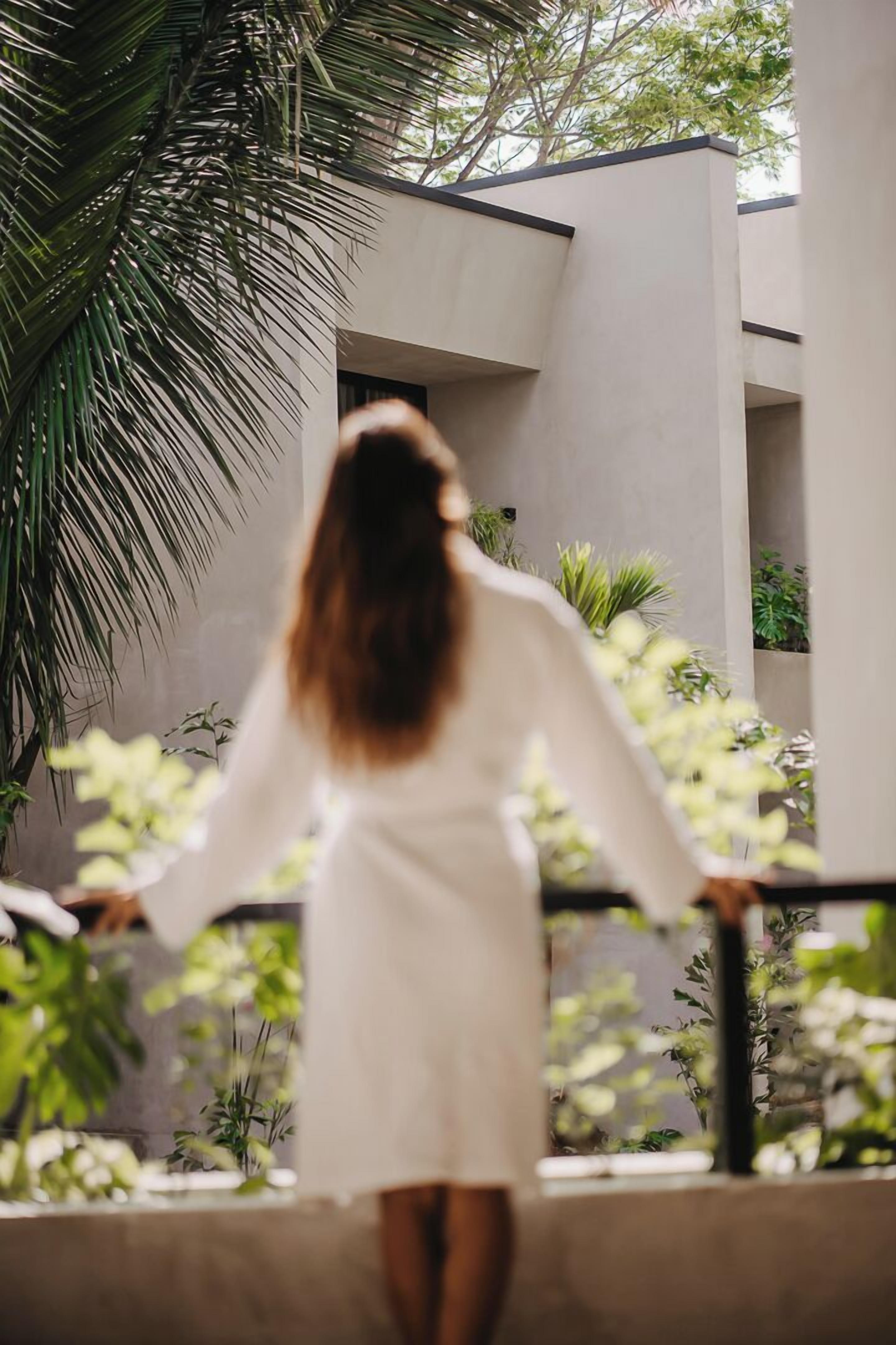 A person in a white dress walking on a balcony with plants and a modern building in the background.