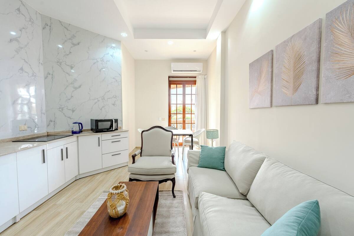 A modern kitchen with white cabinets, a marble backsplash, and a wooden dining table. There is a white sofa and a chair in the living area, and a window with curtains.