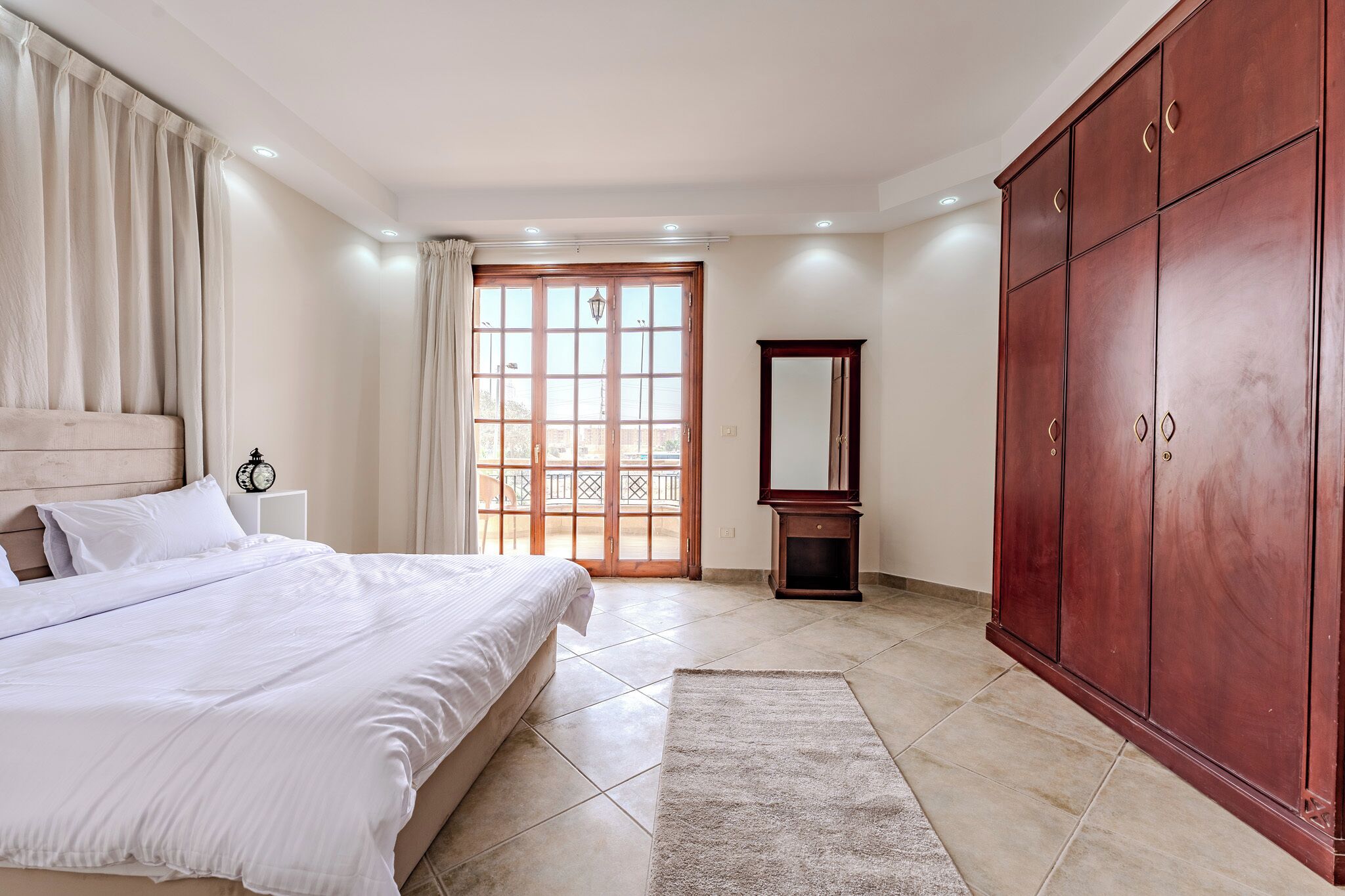 Une chambre à coucher avec un grand lit, une armoire, un miroir et une fenêtre avec des rideaux.