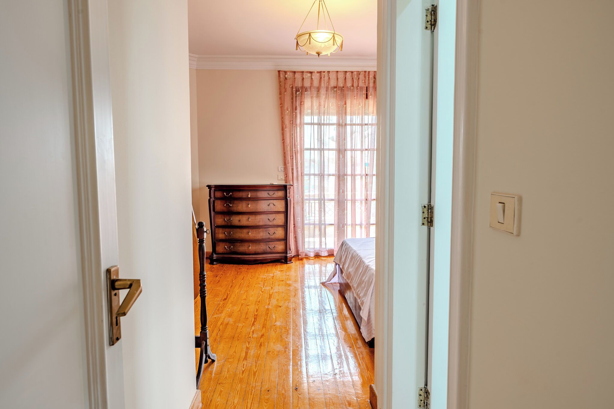 Une pièce avec un plancher en bois, un lit recouvert d’une courtepointe blanche, une commode en bois et une fenêtre avec des rideaux.