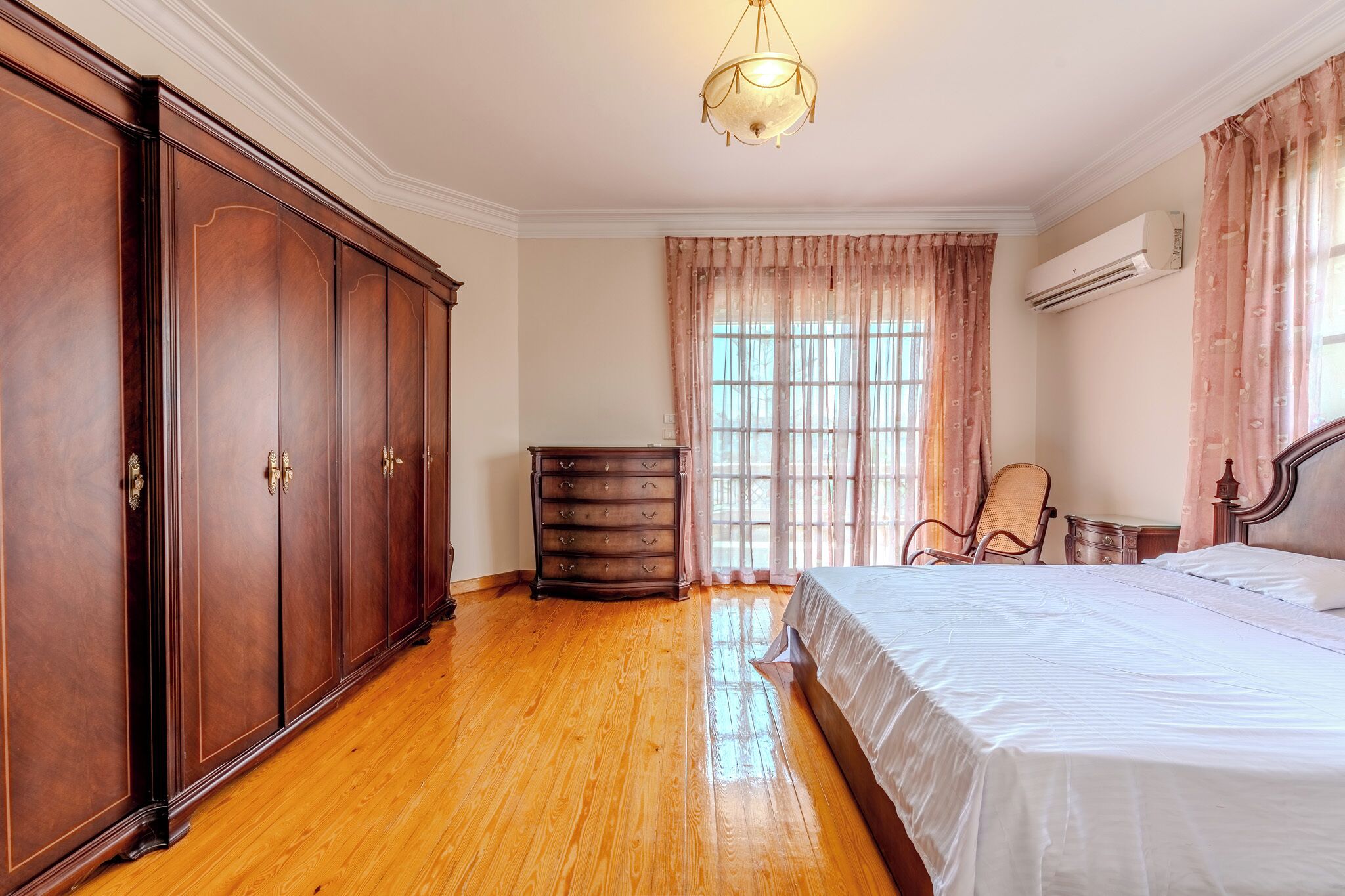 Un dormitorio con un amplio armario de madera, una cama con ropa de cama blanca, una silla y un aparador.