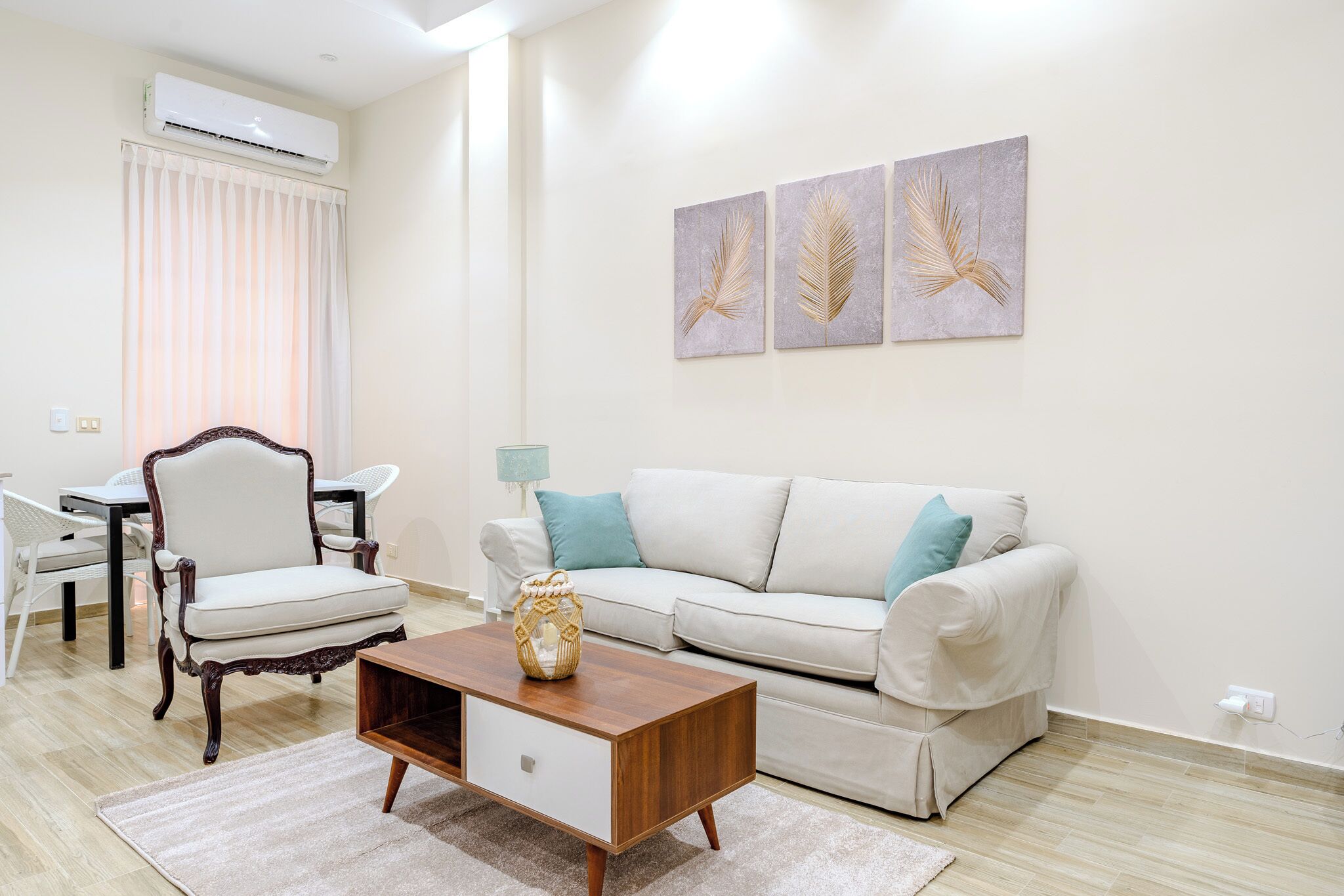 A living room with a white sofa, a wooden coffee table, a decorative vase, and a chair. There is a wall-mounted air conditioner and two wall art pieces.