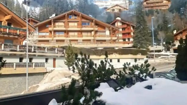 Un bâtiment en bois à plusieurs étages, doté de balcons, avec une rivière au premier plan et un paysage enneigé.