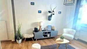 Un salon moderne avec un plancher en bois, un canapé gris, un fauteuil blanc, une petite table basse ronde, un pouf blanc moelleux, un meuble de rangement gris agrémenté d’objets décoratifs et une tête de cerf fixée au mur.