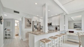 A modern kitchen with white cabinetry, a central island with bar stools, and a dining area with a white chair. - Bay View CLOSE to Beach, Shops, Restaurants, Marina (Bradenton Beach)