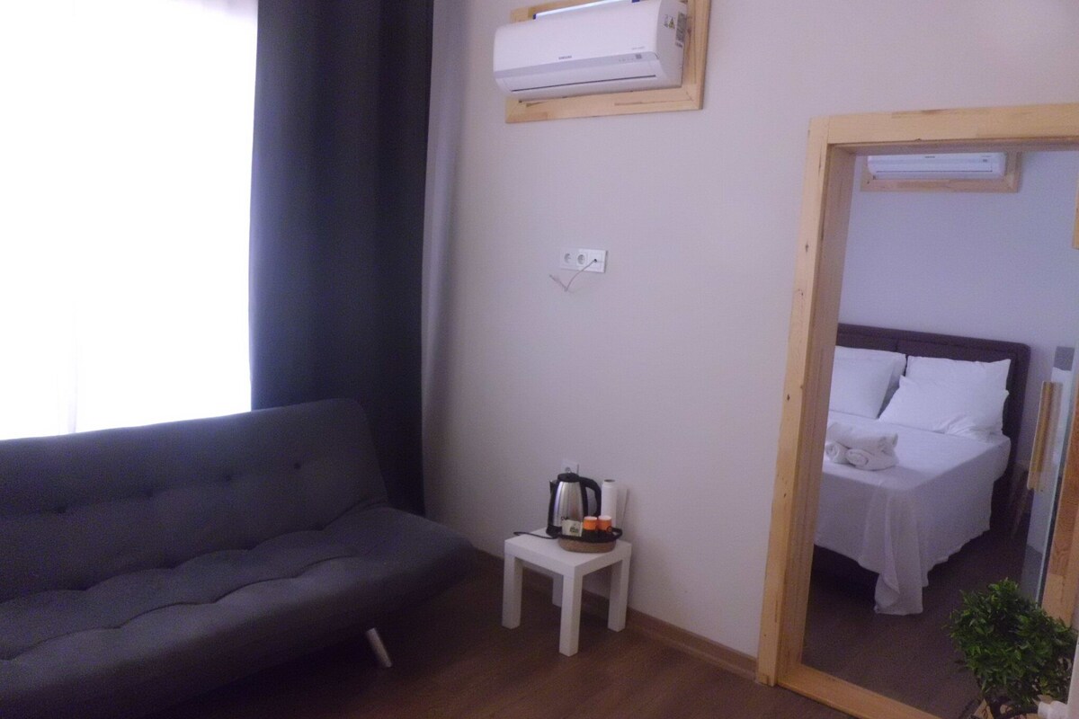 Une pièce avec un canapé gris, une petite table blanche sur laquelle se trouvent une bouilloire et des tasses, et un lit avec du linge de lit blanc.