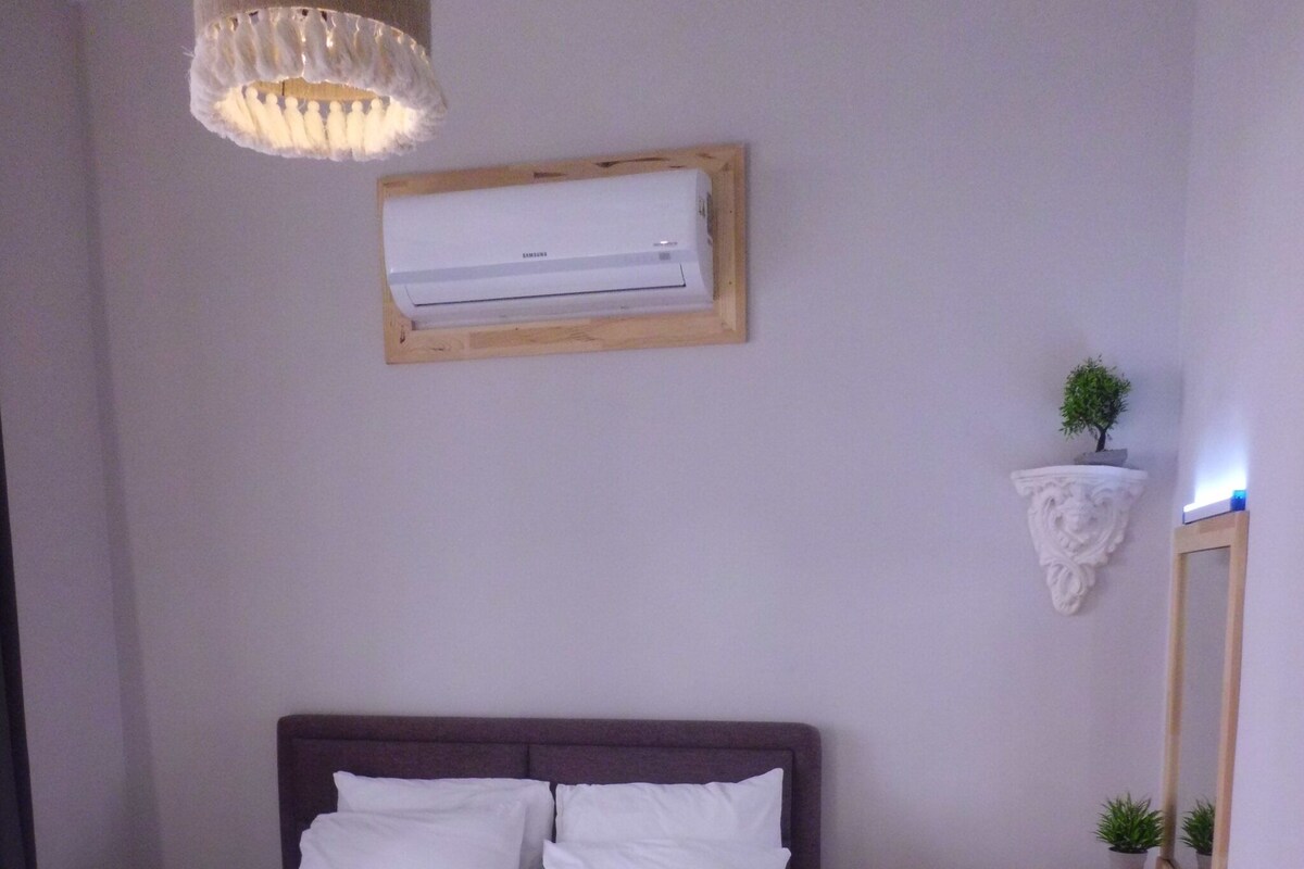 Ein ordentlich bezogenes Bett mit weißen Laken und einem gefalteten Handtuch, ein kleiner Holzschemel, eine Wand-Klimaanlage, ein Spiegel und ein kleiner Topfpflanze auf einem Regal.