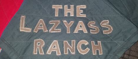 A pillow with embroidered text "THE LAZYASS RANCH" on a green fabric.
