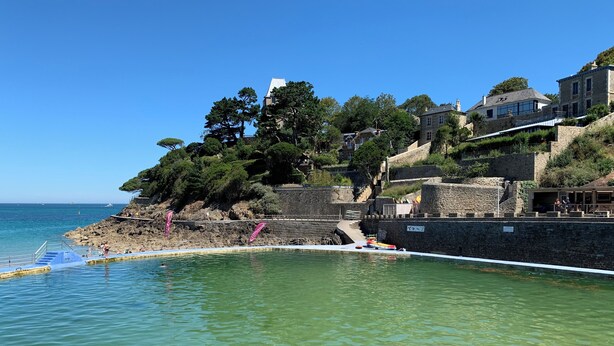 A coastal area with clear water, a stone wall, and buildings on a hillside.