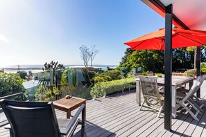A wooden deck with outdoor furniture, including a table and chairs, and a red umbrella providing shade. - 19th Hole Getaway - Mangawhai Holiday Home (Mangawhai)