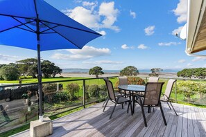 Eine Holzterrasse mit Tisch und Stühlen, mit Blick auf eine Küstenlandschaft mit Bäumen und Meer.