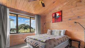 Un dormitorio con techo de madera, un ventanal con cortinas, cama con ropa de cama estampada, un cuadro abstracto rojo y una lámpara de pie.