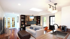 A modern living room with a sofa, ottoman, and a flat-screen TV. There is a kitchen area with a sink and a dining table.