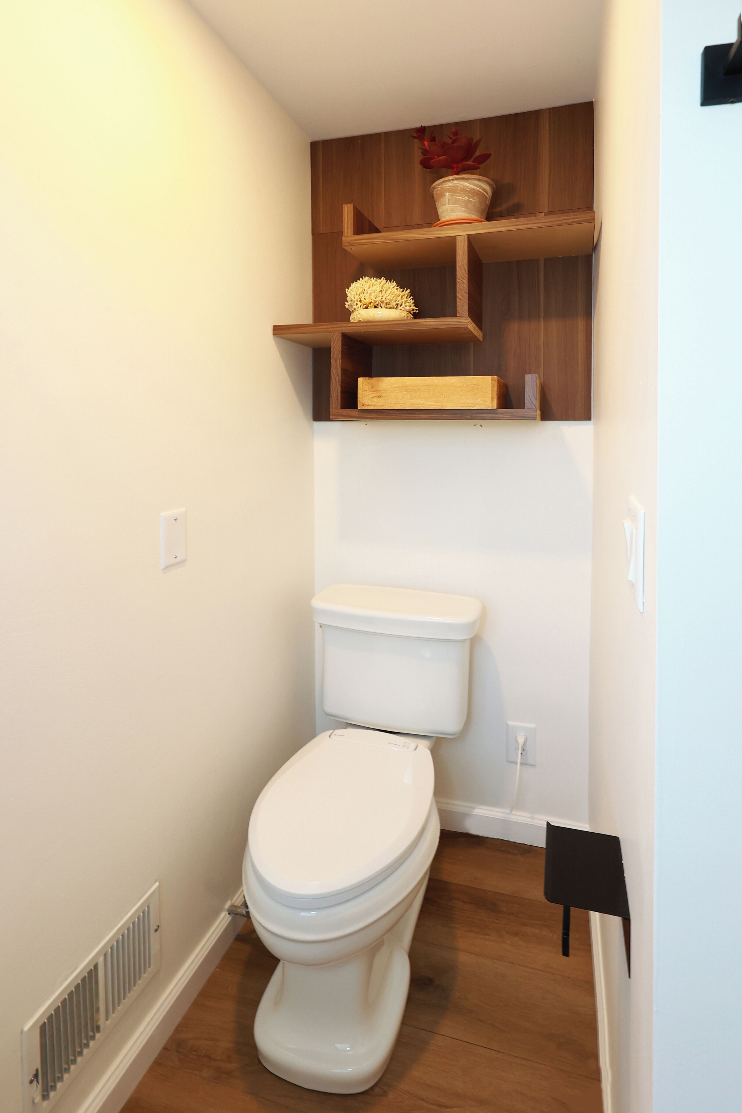 Une petite salle de bain avec des toilettes blanches, des étagères en bois au-dessus et une grille d’aération sur le mur.