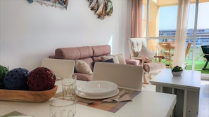 Un salon avec un canapé, une table à manger dressée avec des assiettes, des verres et des couverts, et une vue sur l’extérieur grâce à de grandes fenêtres.