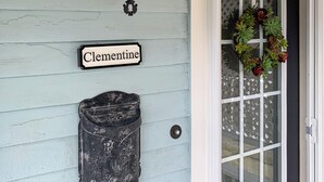 Uma casa com fachada azul clara, um candeeiro preto, uma lata de lixo e uma porta com grinalda.