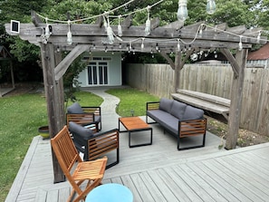 Une terrasse en bois avec des meubles d’extérieur, une pergola illuminée de guirlandes lumineuses et un chemin menant à une maison.