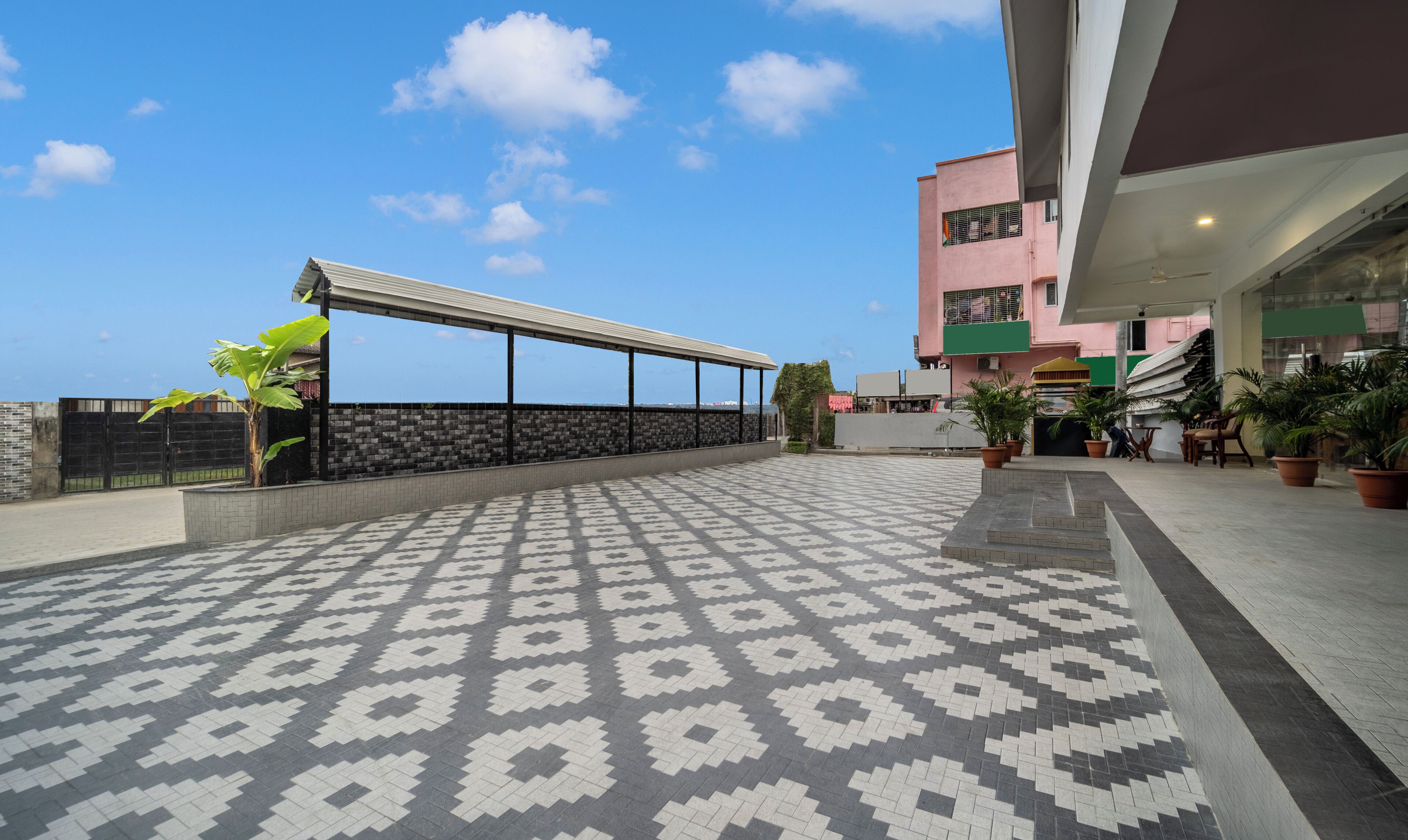 A patterned outdoor area with a covered walkway, potted plants, and a building with a pink facade.