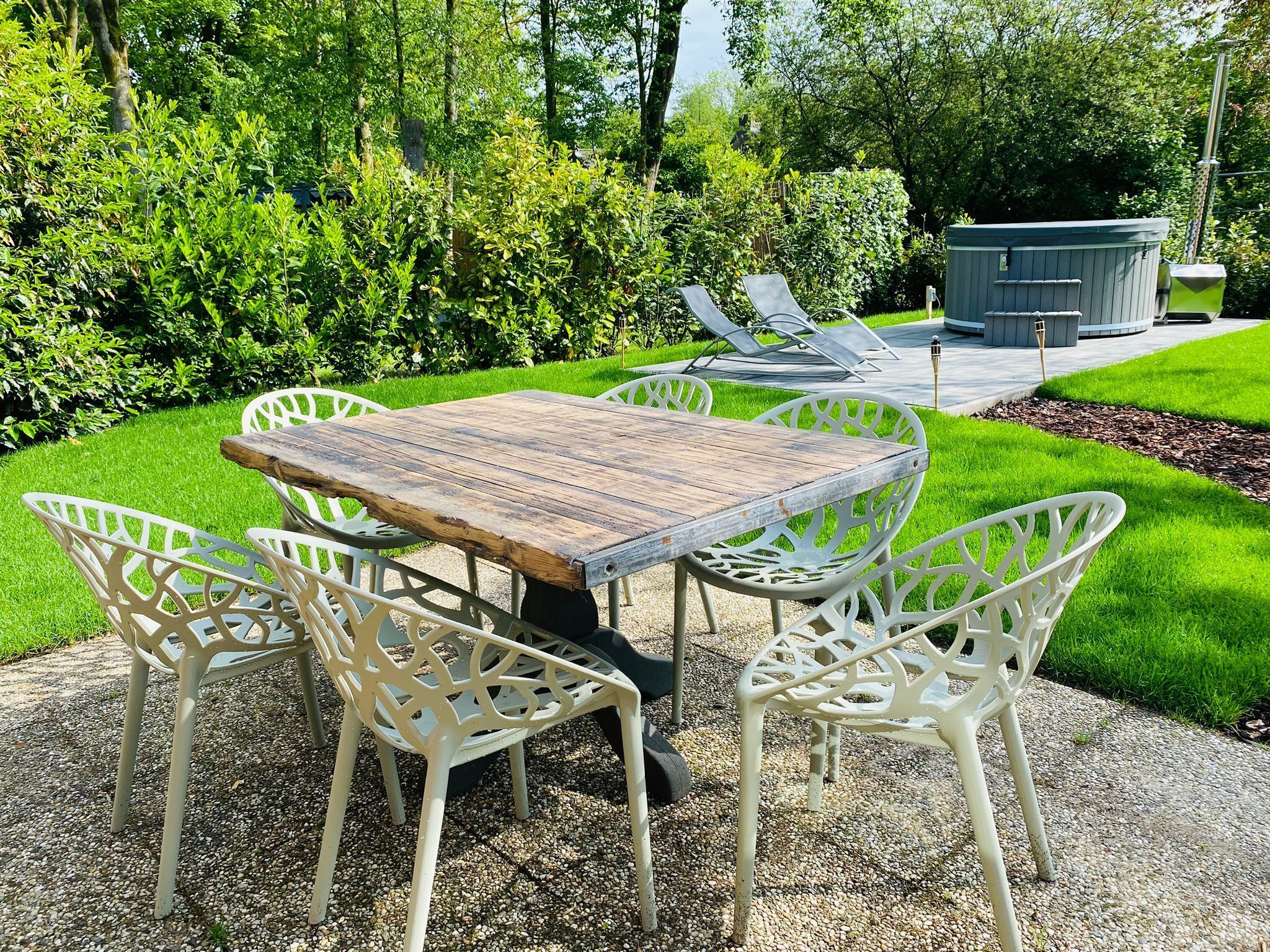 Eine Terrassengruppe im Freien mit einem Holztisch und weißen Metallstühlen, umgeben von üppiger Bepflanzung und einem gepflegten Rasen.