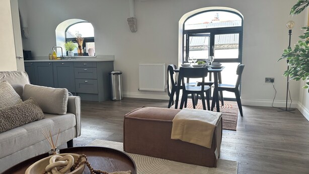 A modern living room with a grey sofa, a brown ottoman, and a dining area with a table and chairs. - Charming 2-bedroom duplex apartment with WiFi in charming Norwich (Norwich)