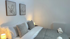 A neatly made bed with white and grey bedding, a grey throw blanket, and a rolled towel on a round ottoman. There are two framed artworks on the wall above the bed. - Charming 2-bedroom duplex apartment with WiFi in charming Norwich (Norwich)