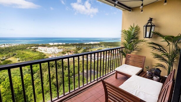 Un balcon avec vue sur l’océan et la verdure, une table en bois, deux chaises et un luminaire suspendu.