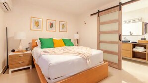 Une chambre à coucher comprenant un lit, des tables de chevet, une lampe, un miroir et une porte coulissante donnant sur une salle de bain.