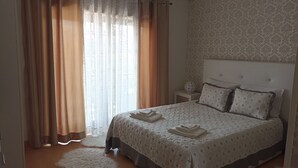 A neatly made bed with a patterned bedspread and pillows, a bedside table with a decorative item, and a window with sheer curtains and a solid curtain partially drawn.