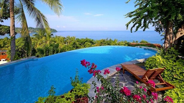 Piscina privada de borde curvo, rodeada de exuberante vegetación y vista al océano.