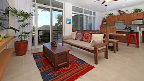 Amplia sala de estar con sofá, mesa de centro y un tapete rojo estampado. Hay un banco de madera, una planta en maceta y una zona de cocina con armarios de madera y un taburete rojo.