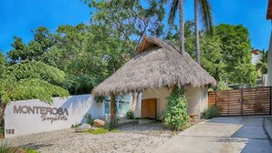 Una casa con techo de paja y un sendero de piedra que conduce a una puerta de madera, rodeada de exuberante vegetación y una palmera.