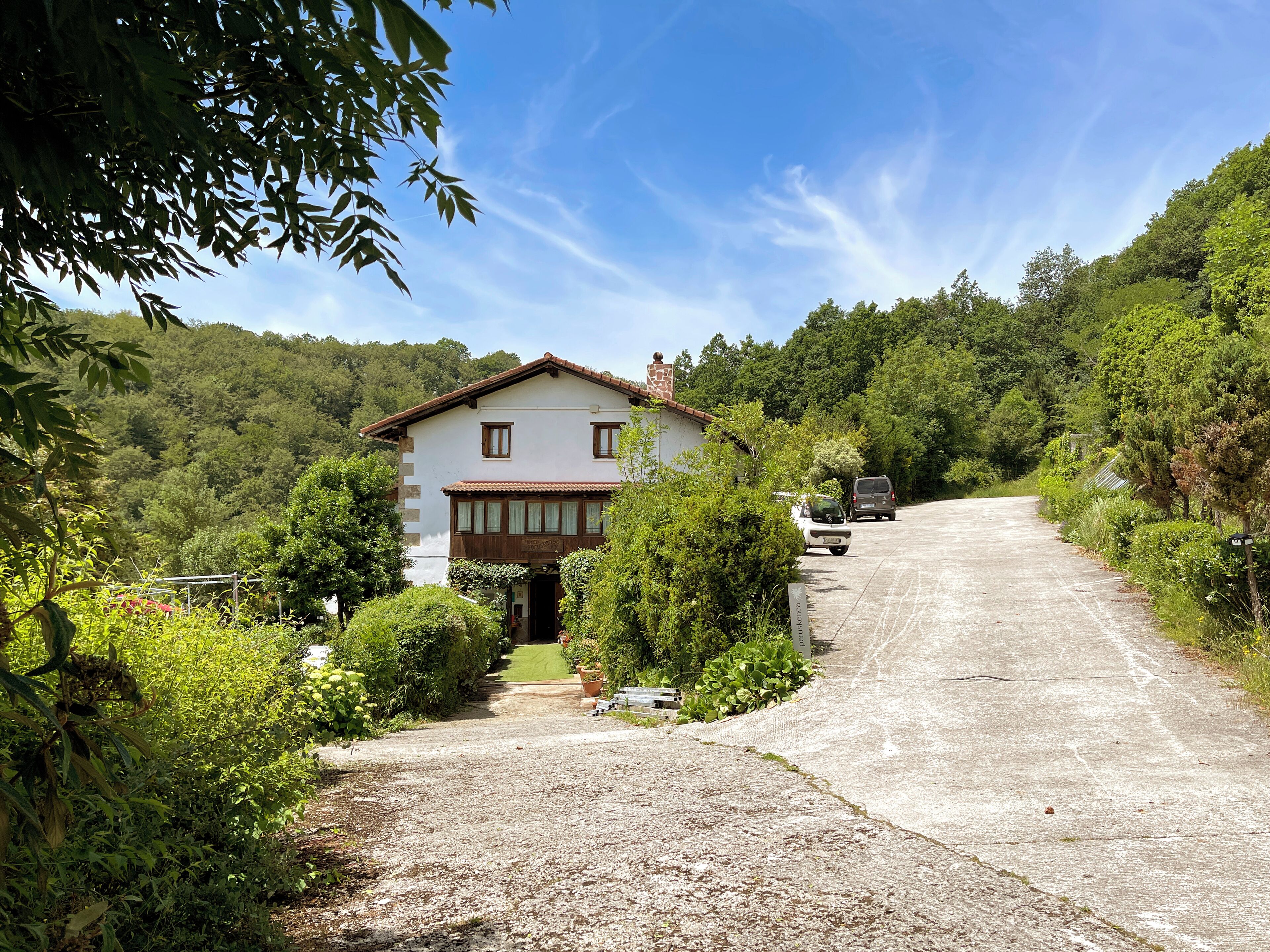 A two-story house with a gravel driveway leading to a wooden door, surrounded by lush greenery and a clear blue sky.