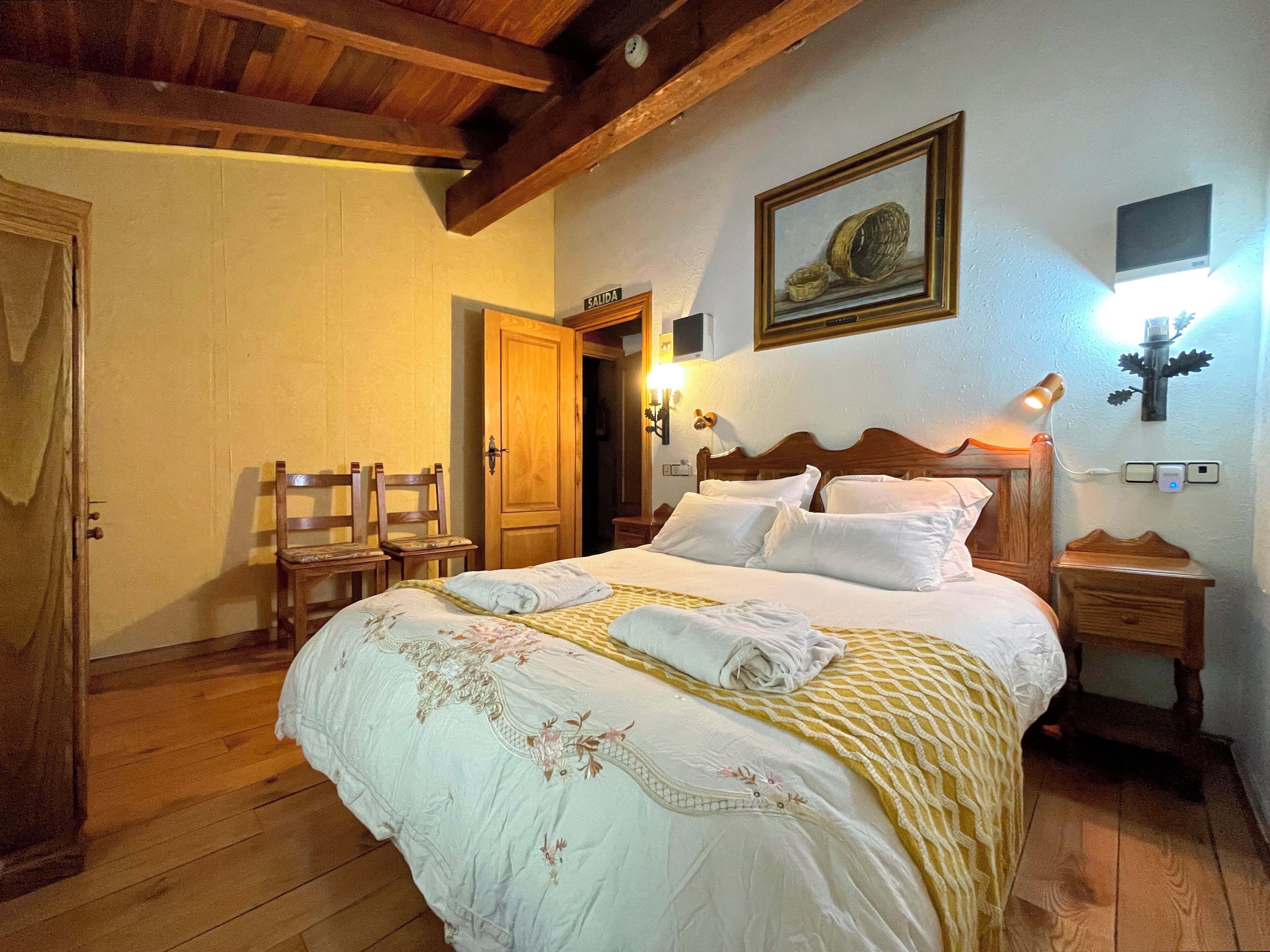 A bedroom with a wooden bed, a nightstand, a bedside table, a lamp, a painting, and a door labeled "SALIDA".
