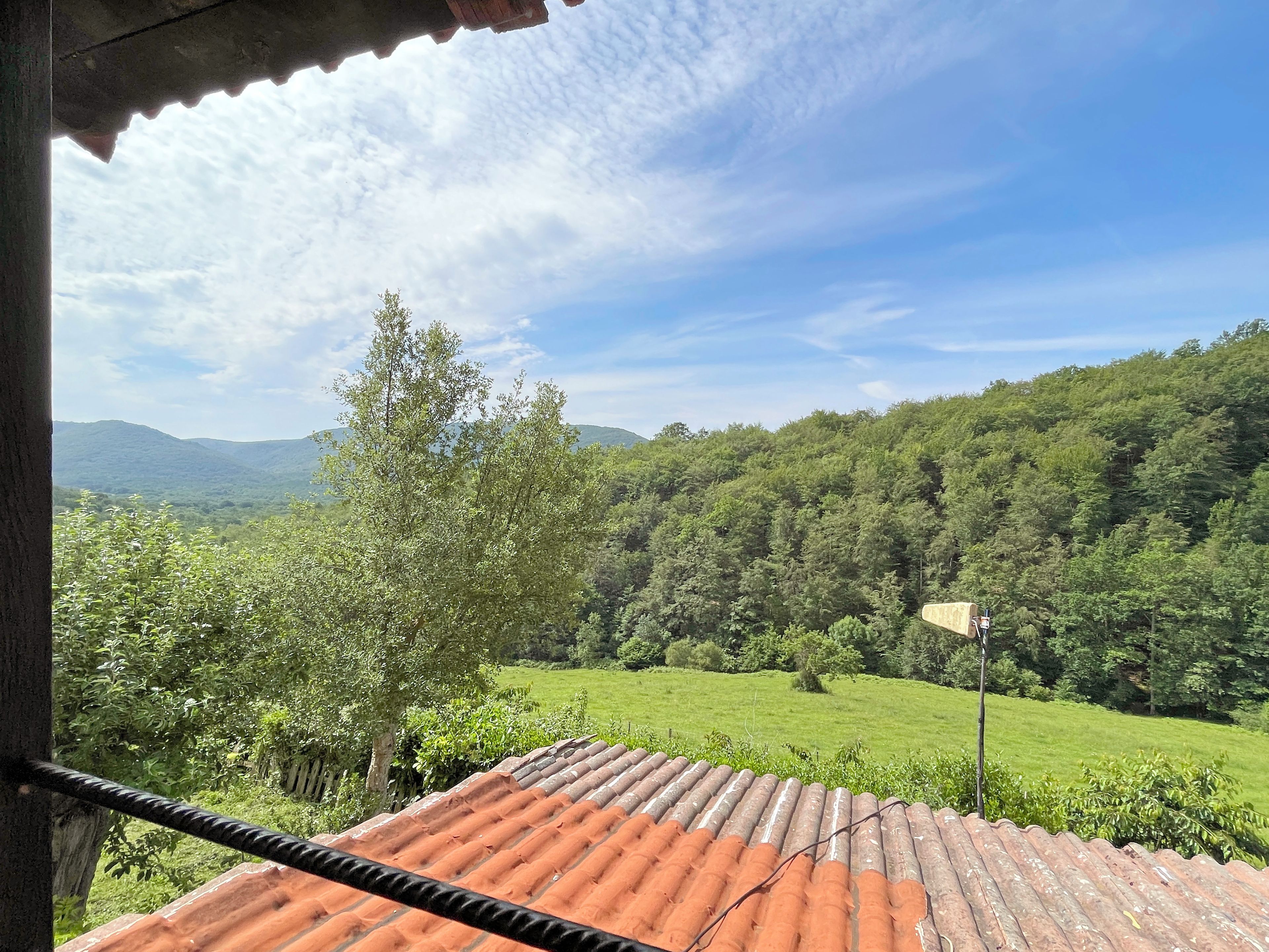 A view from a covered balcony with a red-tiled roof overlooking a lush green landscape and distant mountains.