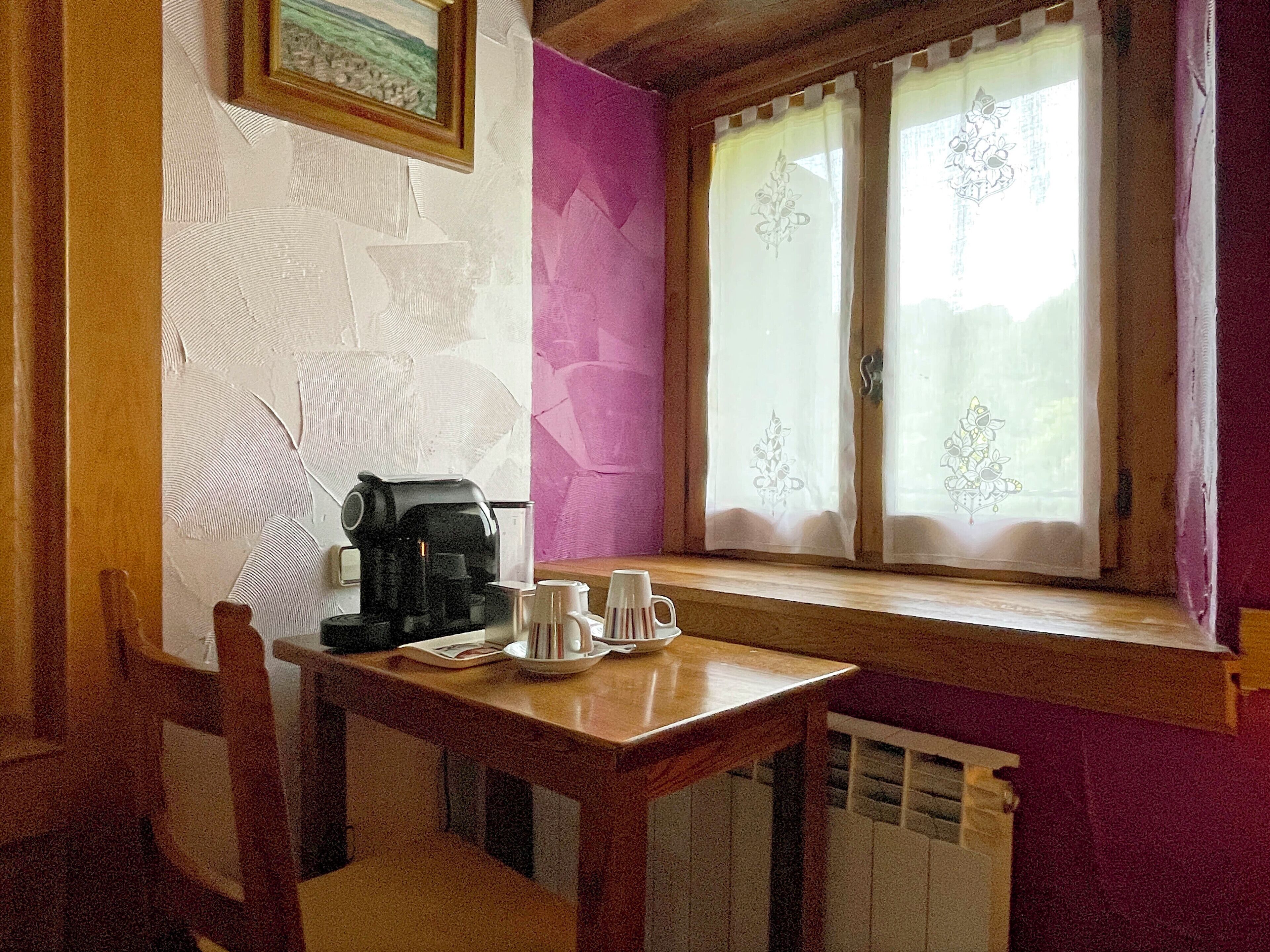 A small kitchen with a coffee maker, cups, and a window with curtains.