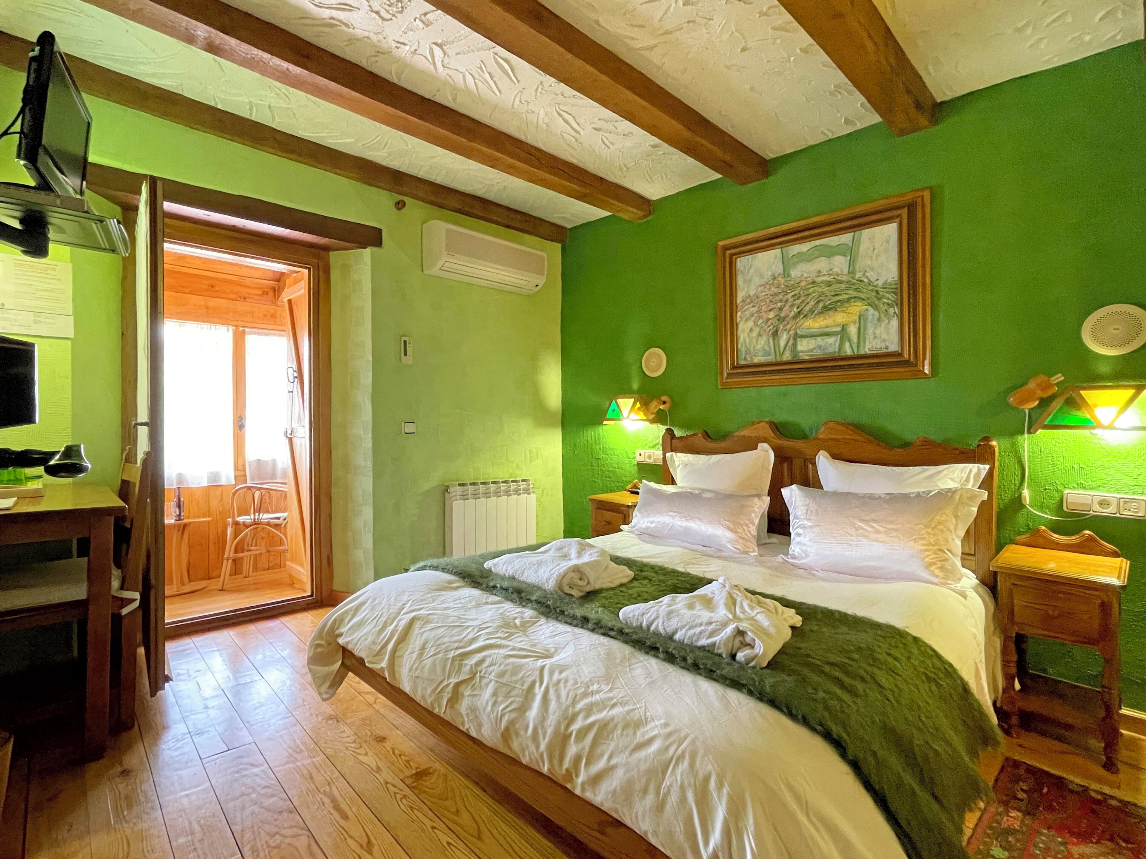 A bedroom with a wooden bed, green walls, and a painting. There is a desk with a computer and a chair.